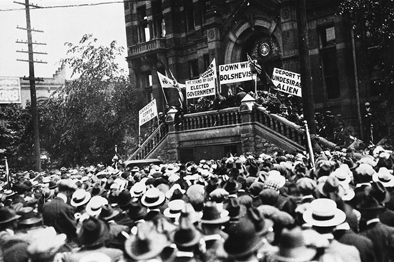 A crowd of people facing away from the camera and looking at people holding signs standing on the stairs in front of a building. 